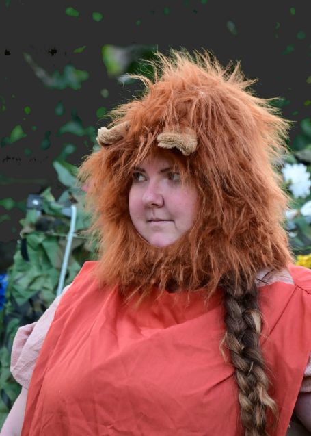 A person wearing a lion costume with a mane and orange clothing, looking thoughtfully.