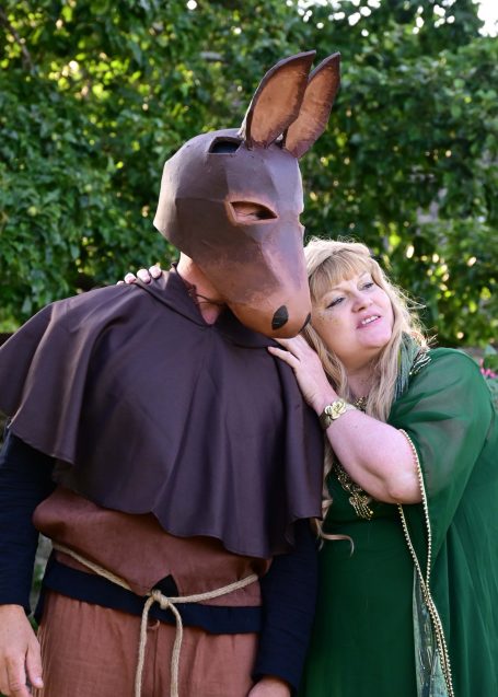 A person in a brown mask resembling a donkey stands beside a woman in green attire.