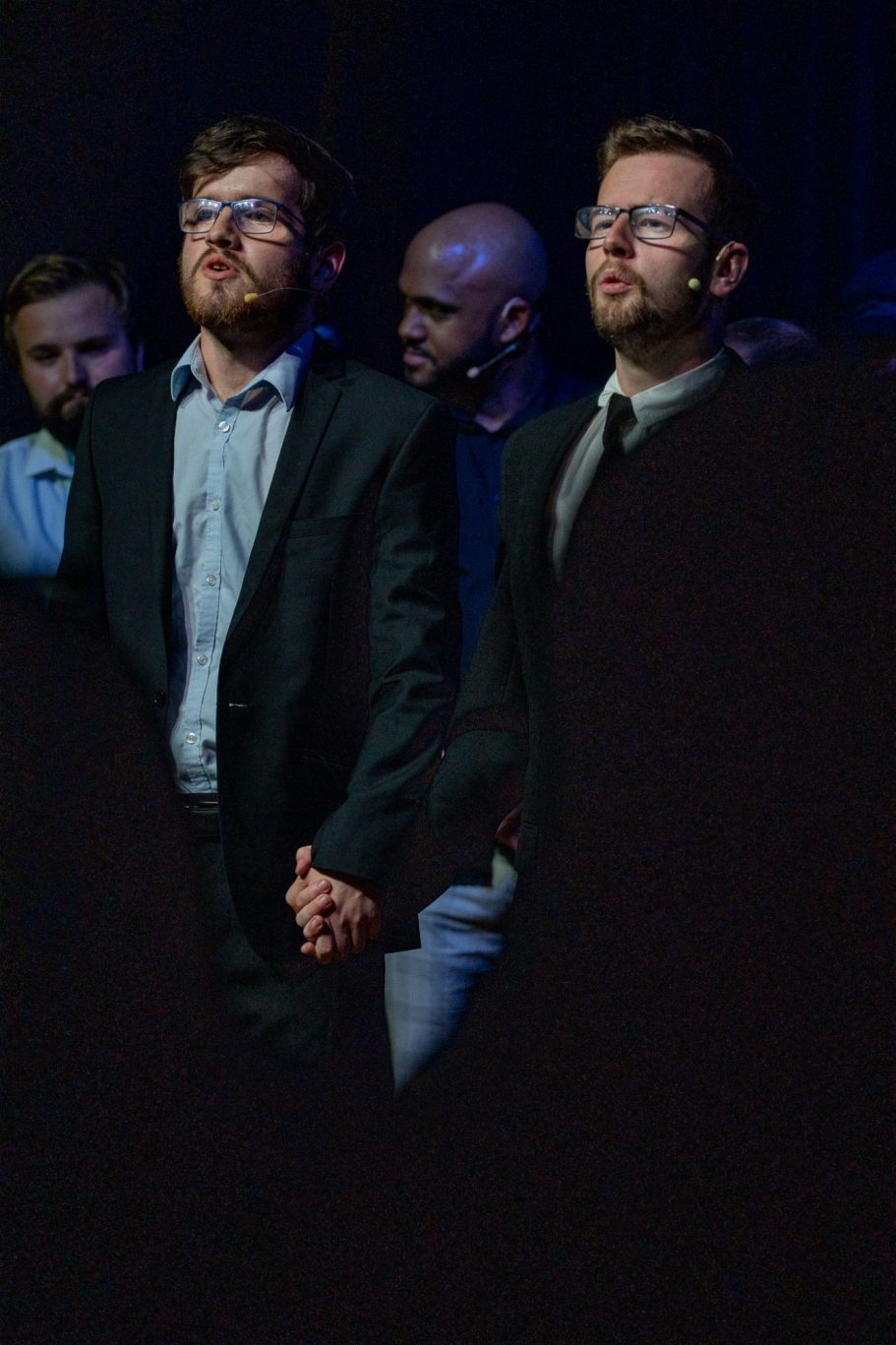 Two men in suits holding hands, standing together on stage.