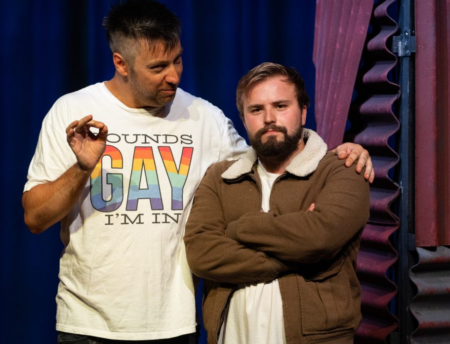 Two men on stage; one wearing a colourful "Sounds Gay I'm In" shirt, the other appears serious.