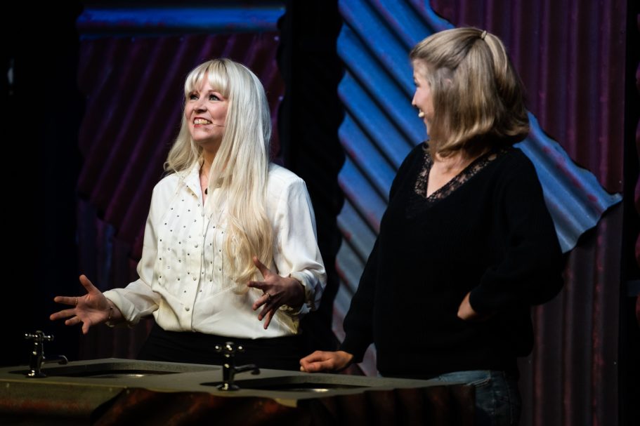 Two women, one with long blonde hair, gesturing during a presentation on stage.