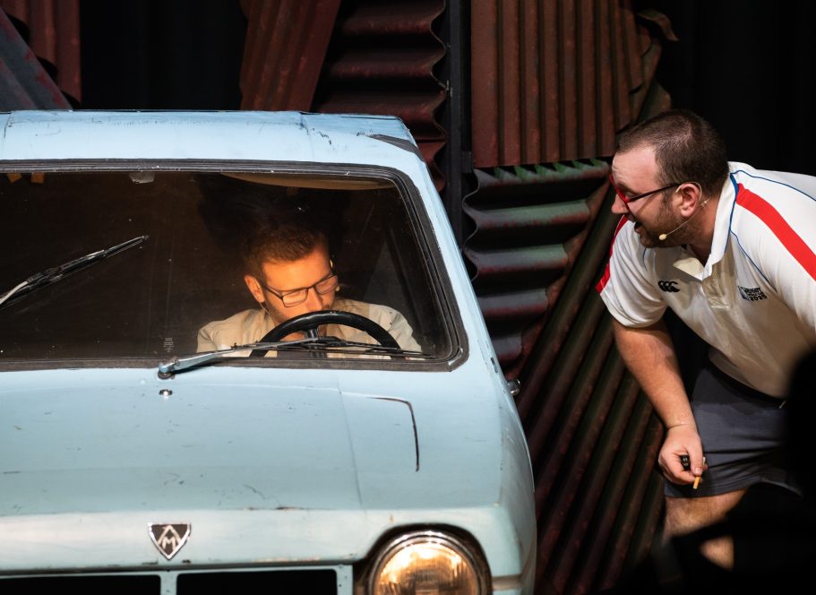 A person sits in a small car while another stands beside, laughing and interacting.