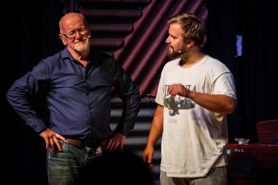 Two men on stage, one smiling and wearing a blue shirt, the other in a white t-shirt.