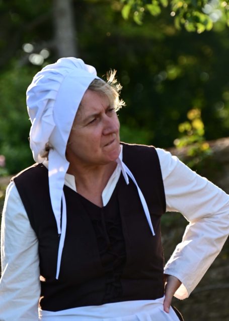 Woman in historical costume with a white bonnet, looking thoughtfully outdoors.