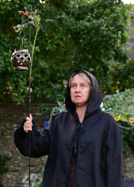 A woman in a black cloak holding a staff adorned with a flower and a decorative pot.