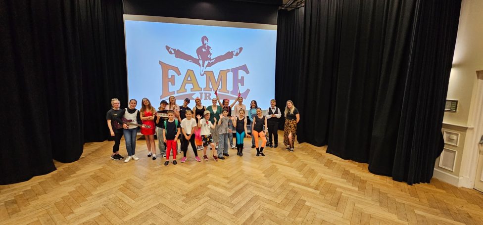 Youth Theatre Show Project Group of children and adults posing together on stage with a "FAME" backdrop.