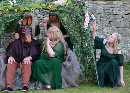 A group in medieval costumes, depicting a scene with greenery and a character in a mask.