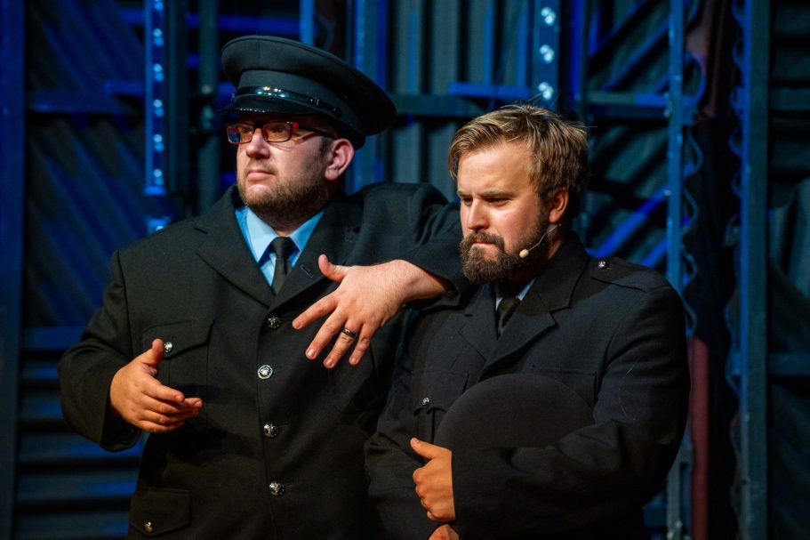 Two men in theatrical costumes, one in a police uniform and the other in a dark suit.