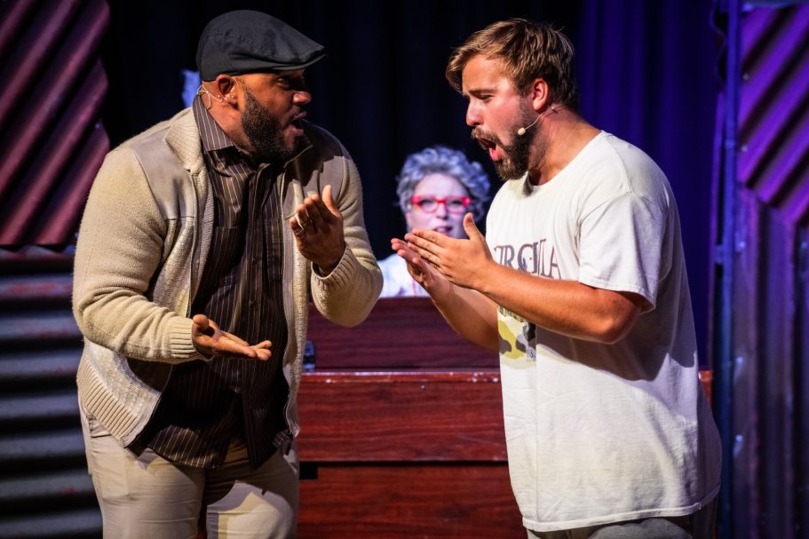 Two men perform animatedly on stage, while a third person observes in the background.