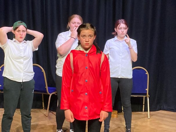Youth Spotlight Drama Crew A girl in a red raincoat stands in front of three peers, gesturing dramatically.