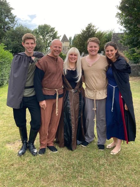 Five people dressed in medieval costumes, posing together outdoors on a grassy area.