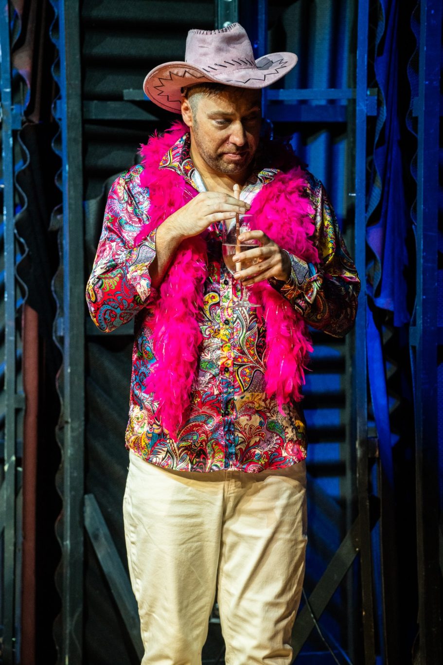 A man wearing a colourful outfit, pink feather boa, and cowboy hat, standing on stage.