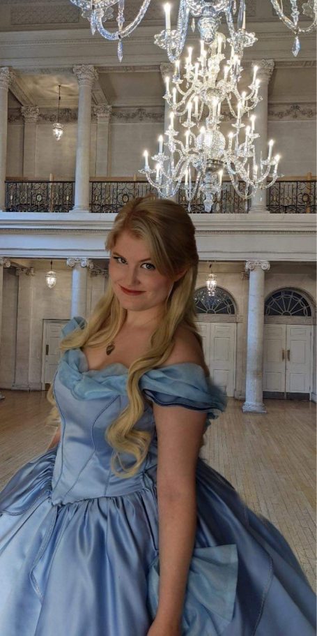 Young woman in a light blue gown with long blonde hair, smiling in a grand room.