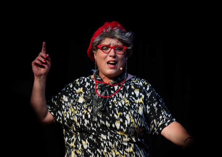 A performer with glasses and a red hat gestures expressively on stage.