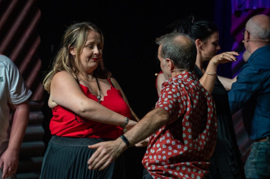 Two people dance closely on stage, surrounded by others in a lively performance.
