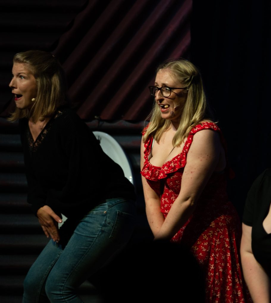 Two women perform on stage, one in a black top and the other in a red dress.