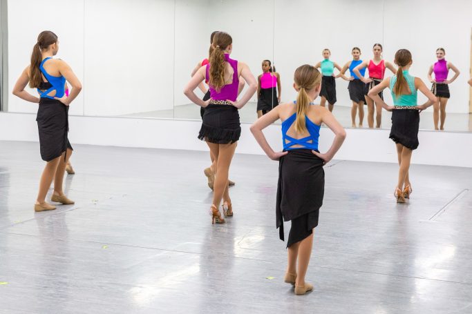 BallroomKids Group of dancers in bright tops and black skirts practising in a mirrored studio.