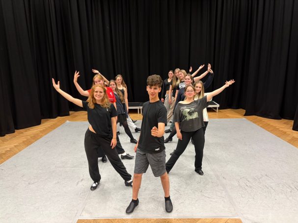 Beginners Tap (Adults and Teens) A group of dancers posing on stage with arms raised, showcasing a dynamic performance.