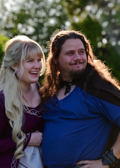 Two people dressed in costumes, smiling outdoors with greenery in the background.