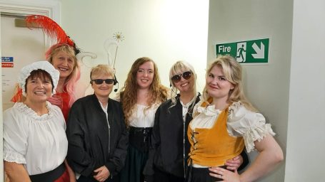 Group of six people in costumes, smiling, posing together in a well-lit hallway.