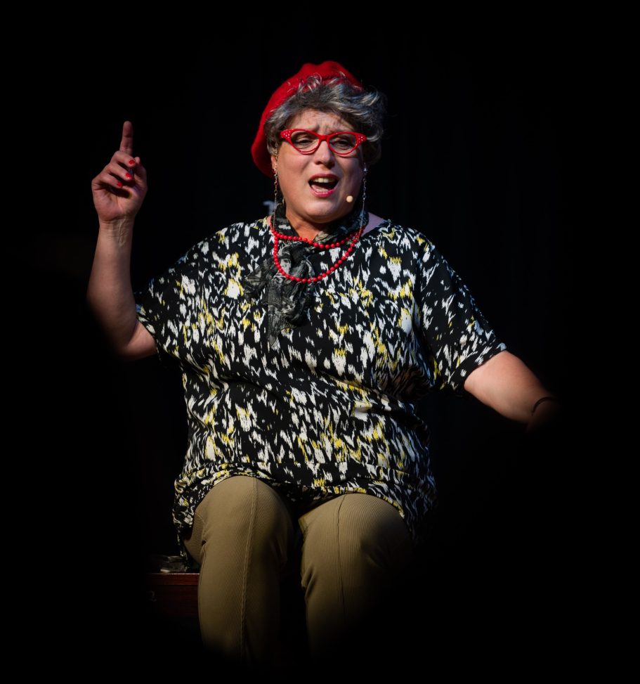 A performer in vibrant clothing gestures expressively while seated on stage.