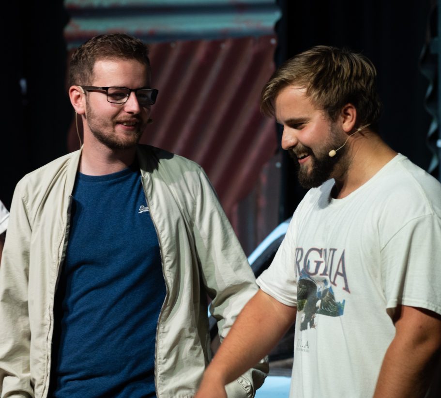 Two men on stage, engaged in conversation, one wearing glasses and a light jacket.