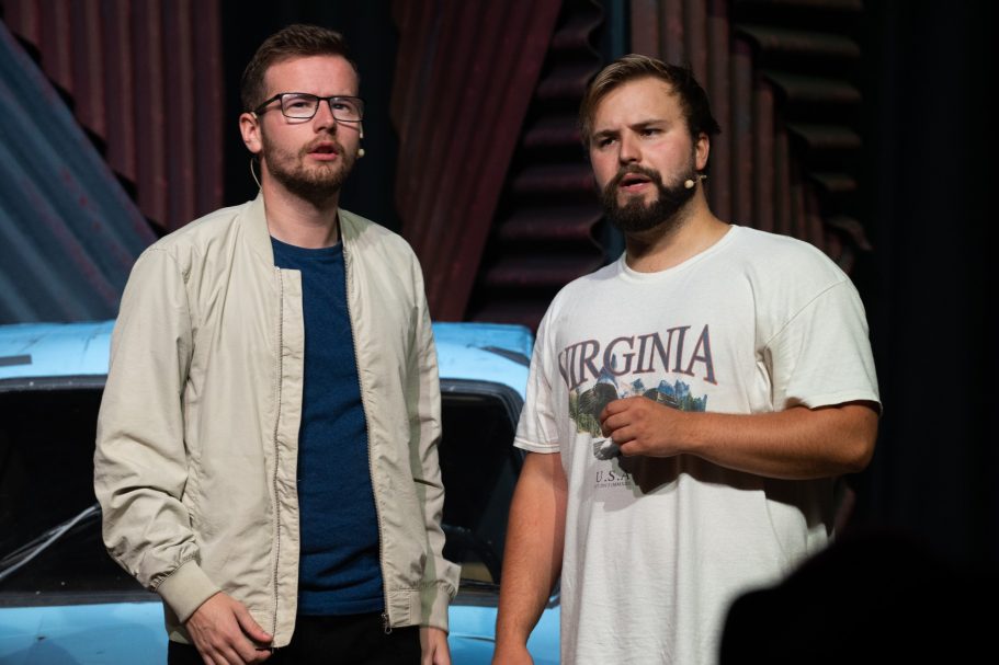 Two men on stage, one wearing glasses and a jacket, the other in a t-shirt.